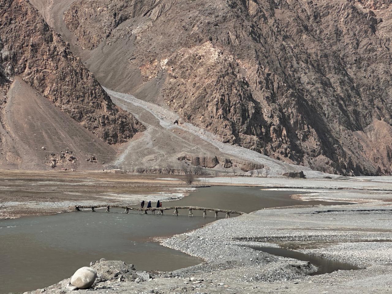 A community crosses a makeshift bridge over a river with mountains behind — the remote terrain Adam Holdings serves