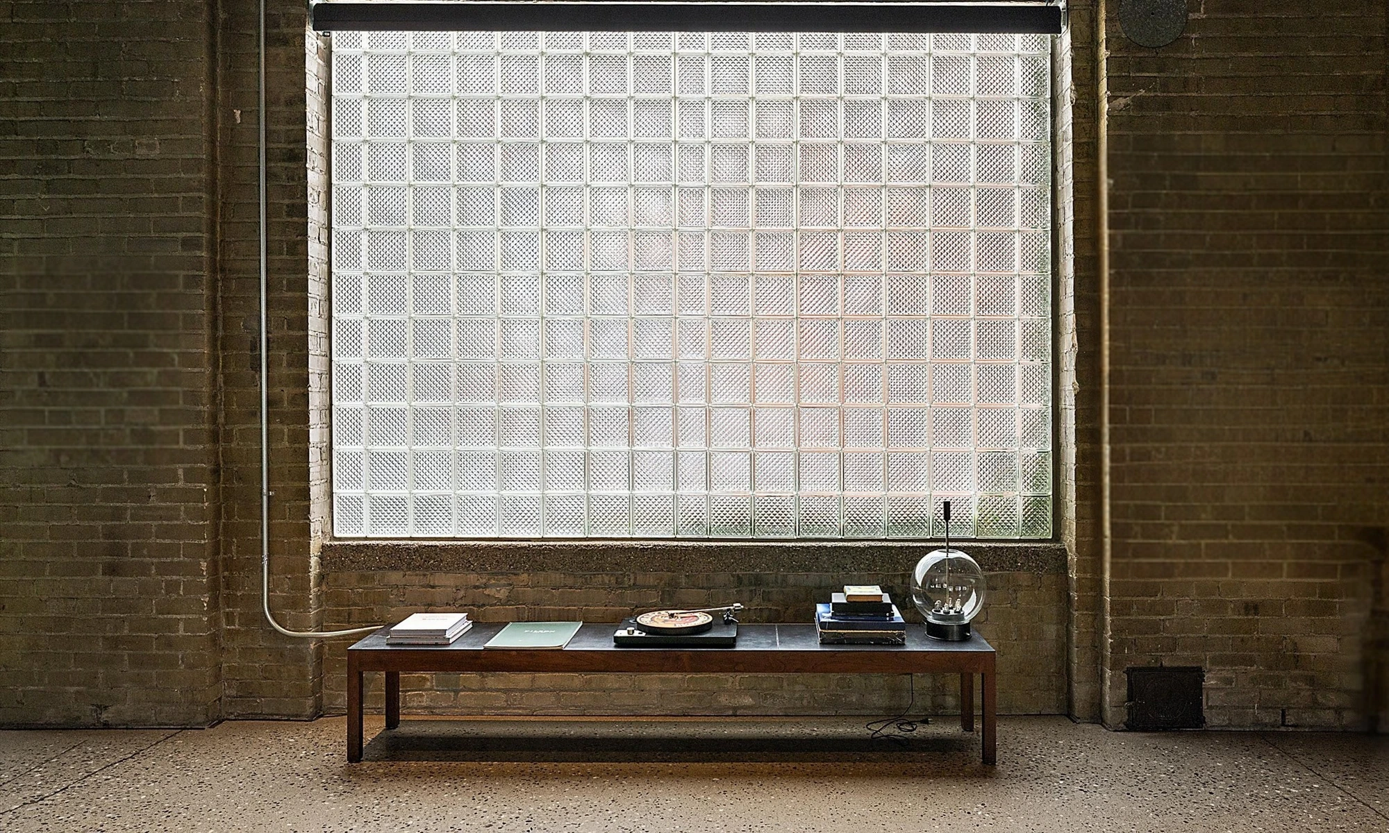 Shop Dog, long dark walnut console beneath glass-block window, turntable, curated books and globe lamp against exposed brick